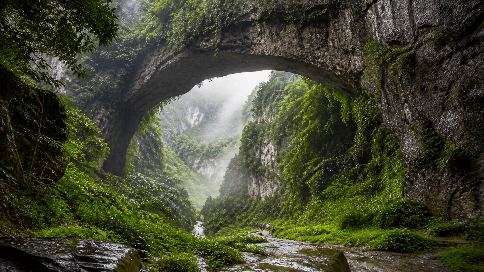 Misty limestone arch and small visitors on the Wulong Three Natural Bridges valley floor