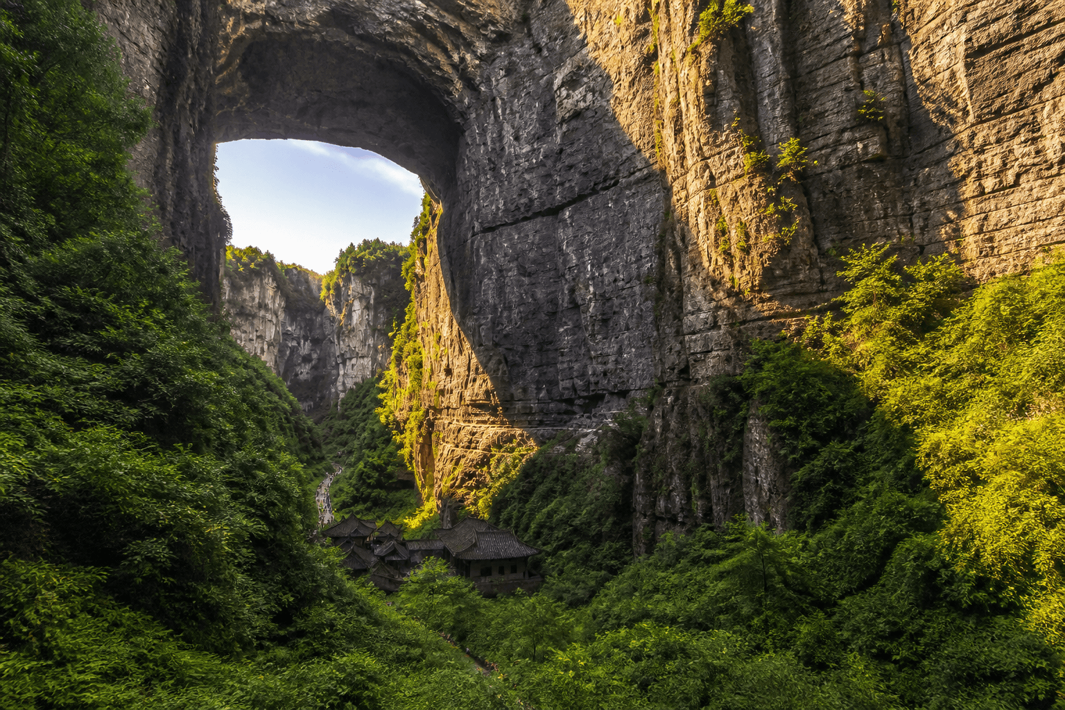 Traditional-style building beneath a large natural arch, with distant visitors for scale